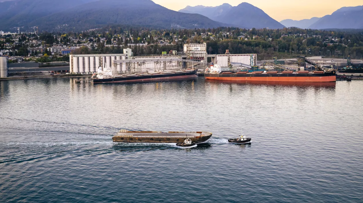 A tug boat pulls a barge through the water. A port terminal can be seen in the background.