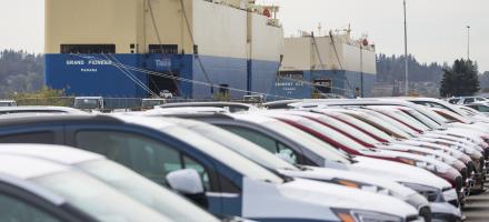 Une rangée de voitures dans un parking. Un navire roulier est visible à l'arrière-plan.